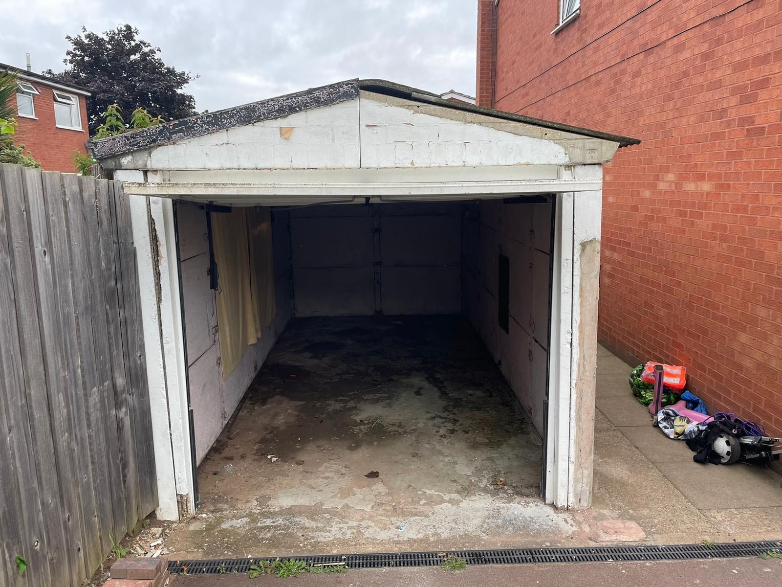A concrete pre-fab garage with an asbestos roof on a driveway , with a fence to the left hand side