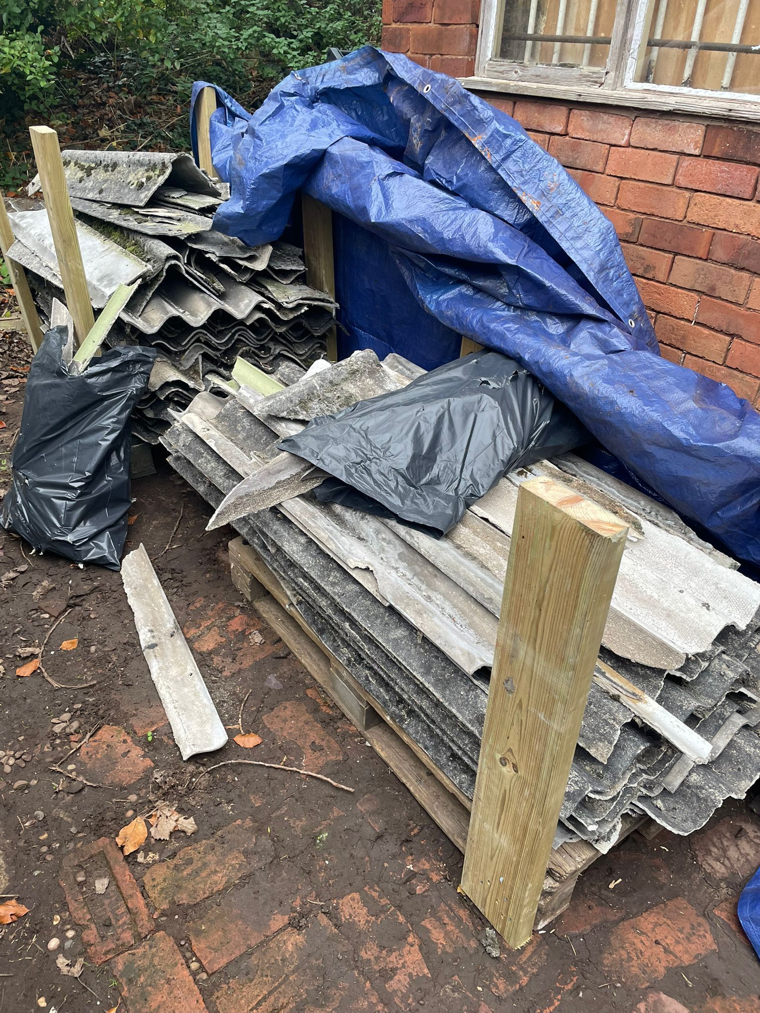 a pile of asbestos cement sheets with a blue plastic sheet , next to a house