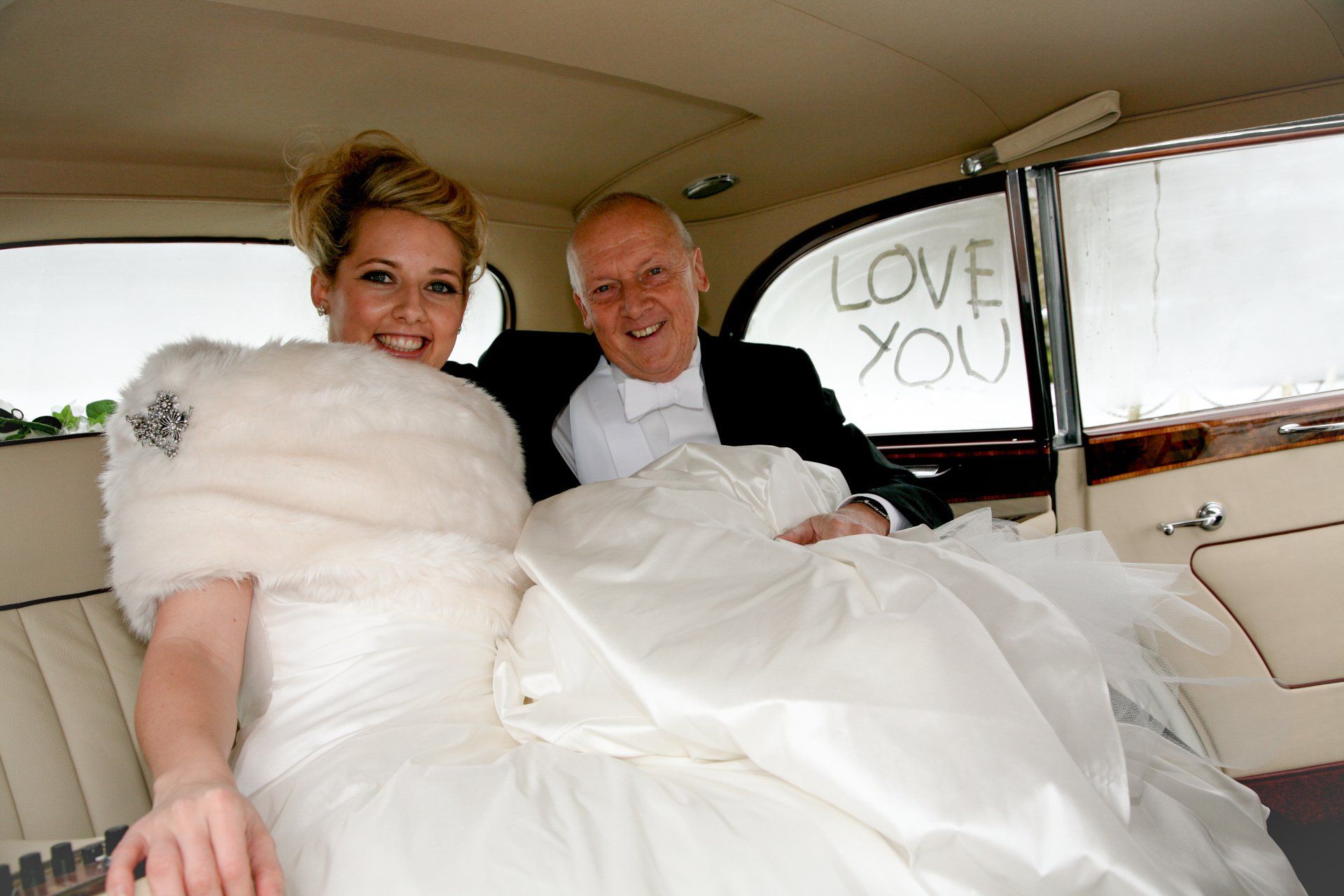 bride in wedding car with her pather and both smiling at the camera