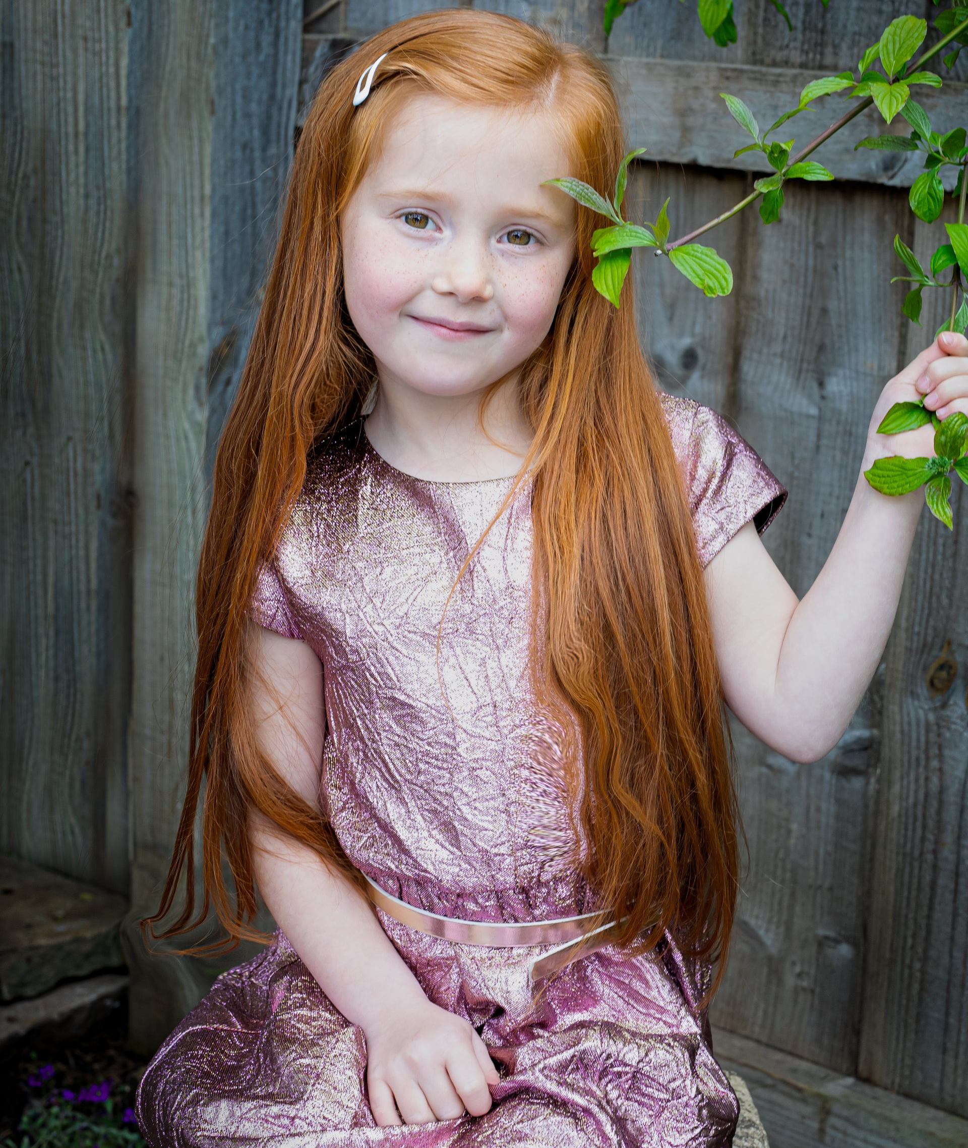 little girl with long ginger hair holding a branch looking at the camera with a little smile
