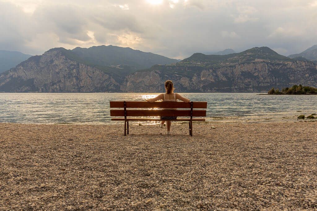 Schöne Momente beim Solo-Travel Eine allein reisende Frau sitzt auf der Bank und blickt auf den Gardasee, im Hintergrund sind Berge.