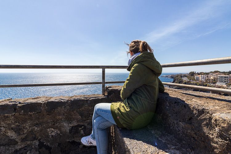 Kurze Pause beim Besuch des Aci Castello. Eine Frau im grünen Parka blickt von der Burg Aci Castello auf die Küste.