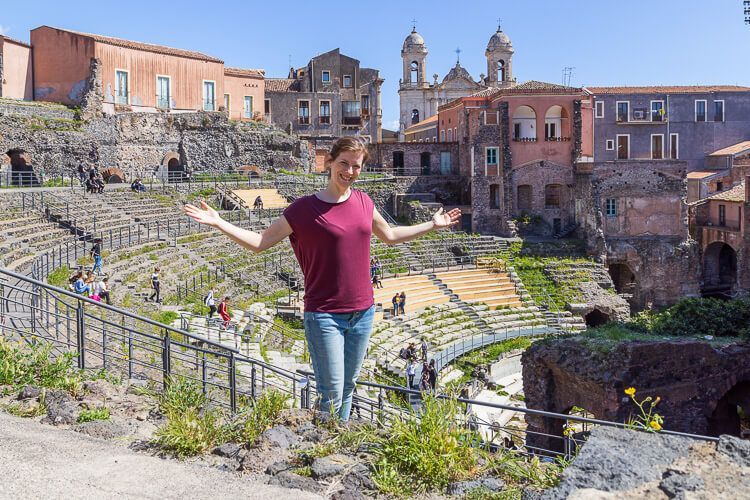 Das Römische Theater in Catania mit bunten Fassaden. Eine lachende Frau steht vor der Kulisse des antiken Römischen Theaters in Catania.