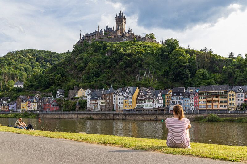 Den besten Blick auf Cochem gibt es vom Stadtteil Cond. Eine Frau sitzt an der Moselpromenade und blickt auf die Altstadt und die Burg von Cochem.
