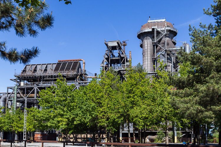 Natur auf dem alten Industriegelände. Hochöfen im Landschaftspark Duisburg-Nord ragen hinter grünen Bäumen hervor.