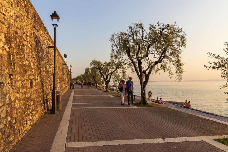 Seepromenade bei Garda kurz vor Sonnenuntergang. Uferpromenade bei Garda bei schönen Abendlicht. Der Weg ist gesäumt von Olivenbäumen.