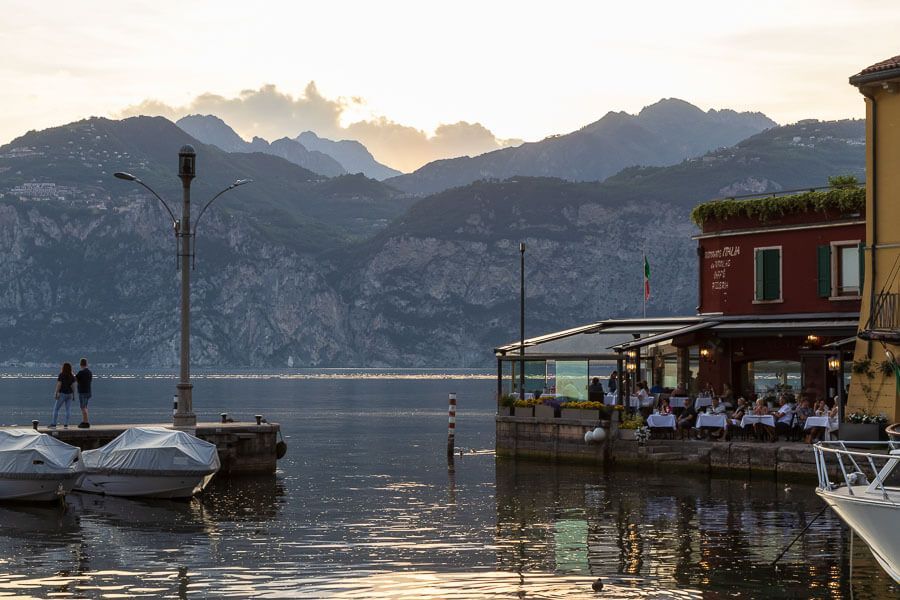Abendstimmung am Hafen in der Altstadt von Malcesine am Gardasee. Hafen von Malcesine mit Blick auf den Gardasee, ein Restaurant und das gegenüberliegende Ufer bei Sonnenuntergang.