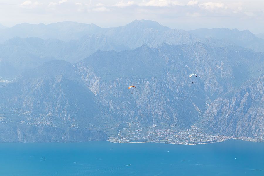 Paraglider, die vom Monte Baldo aus gestartet sind. Paraglider fliegen über dem Gardasee, im Hintergrund sind beeindruckende Bergpanoramen zu sehen.