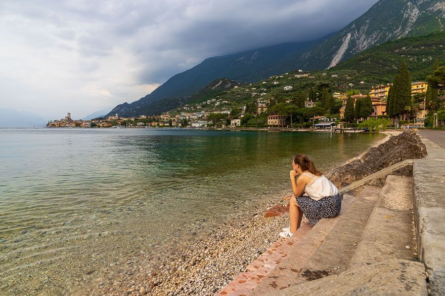 Ausblick auf Malcesine genießen. Eine Frau sitzt am Ufer des Gardasees südlich von Malcesine mit Blick auf eine satt-grüne Bergkette.