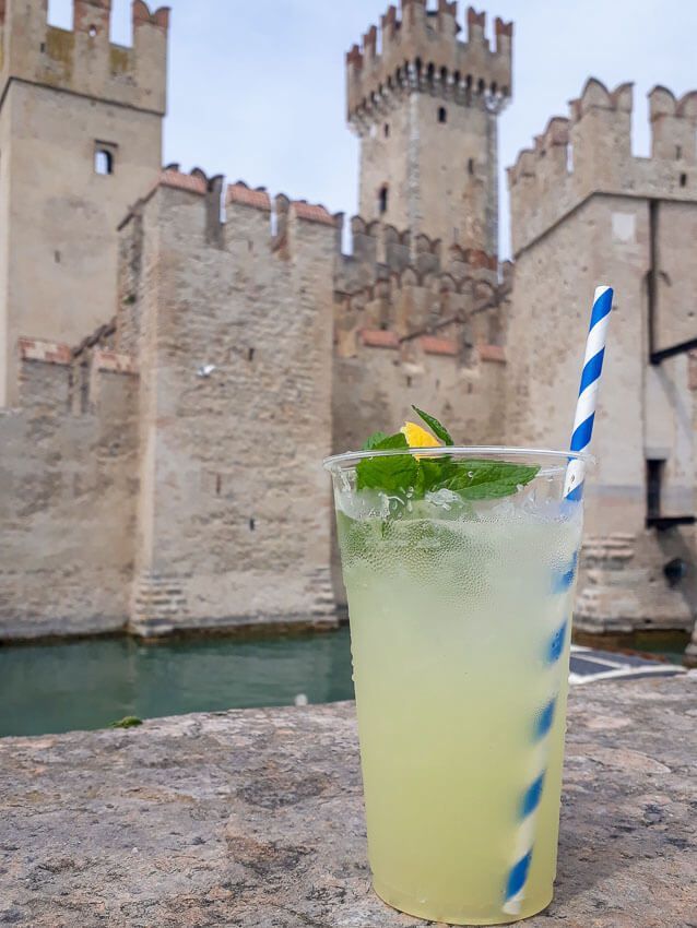 Eine frische Limonade mit blau-weißem Strohhalm. Im Hintergrund thront die Scaligerburg von Sirmione