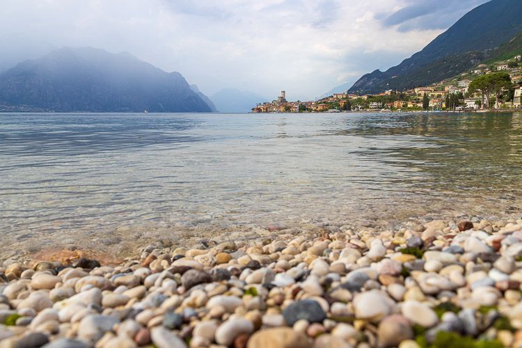 Blick auf Malcesine von einer gegenüberliegenden Bucht. Blick auf Malcesine von einer gegenüberliegenden Bucht. Im Vordergrund sind Kieselsteine des Ufers vom Gardasee.