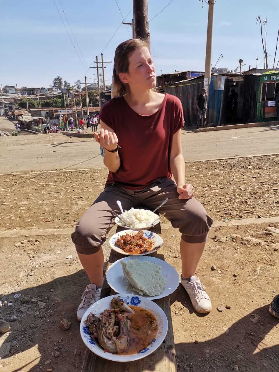 Erste Station der Slumtour. Hier wurden meine Guides und ich von einem der Anwohner mit reichlich Essen versorgt Auf einer Anhöhe mit Blick übers Township. Frau sitzt auf einer Bank mit einer Auswahl an kenianischen Essen