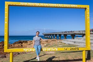 Eine Frau sitzt in einem gelben Panorama-Bilderrahmen am Strand Gqeberha und lässt sich fotografieren.