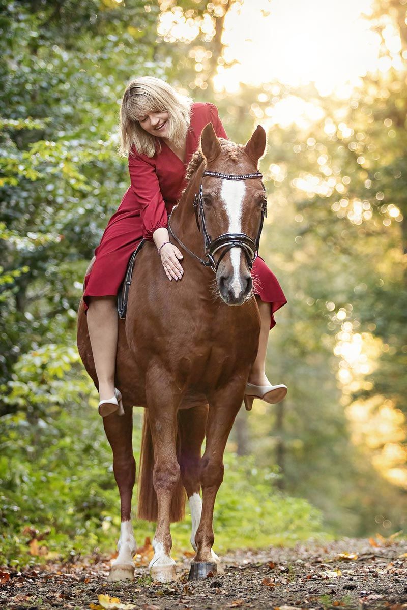LL Photography Mensch und Tier: blonde Frau in rotem Kleid auf einem fuchsfarbenen Deutschen Reitpony in einem lichtdurchfluteten Wald.