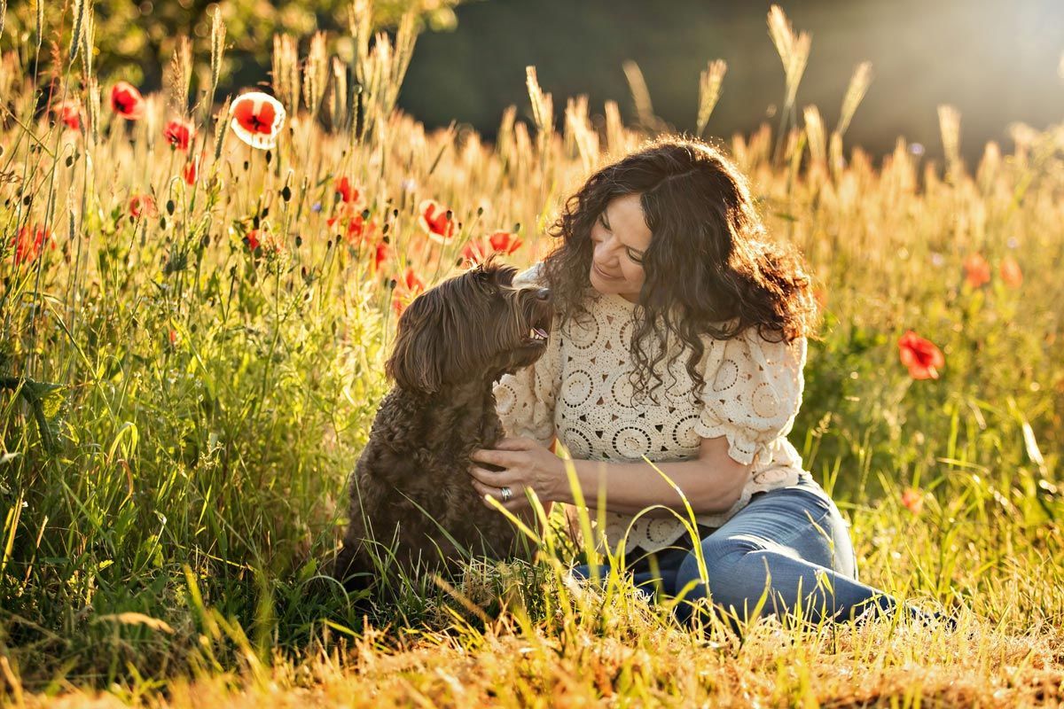 LL Photography Mensch und Tier: eine Frau und ein brauner Labradoodle sitzen vor einem Mohnfeld in der Morgensonne und schauen sich liebevoll an.