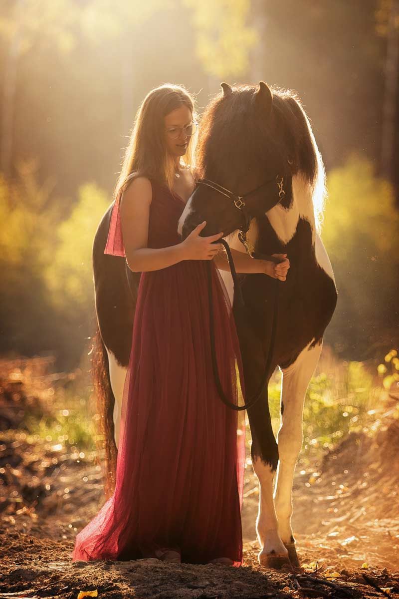LL Photography Mensch und Tier: schwarz-weißer Isländer mit junger Frau in langem roten Kleid im Morgenlicht im Wald.