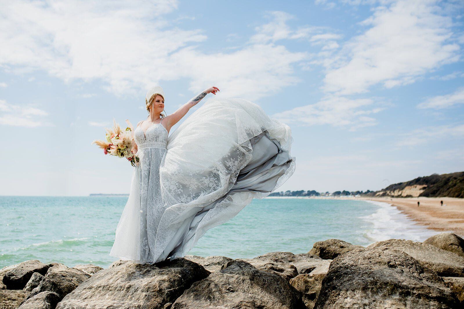 Bride stood on rocks on groyne throwing the train of her dress in the air on Highcliffe Beach in Dorset