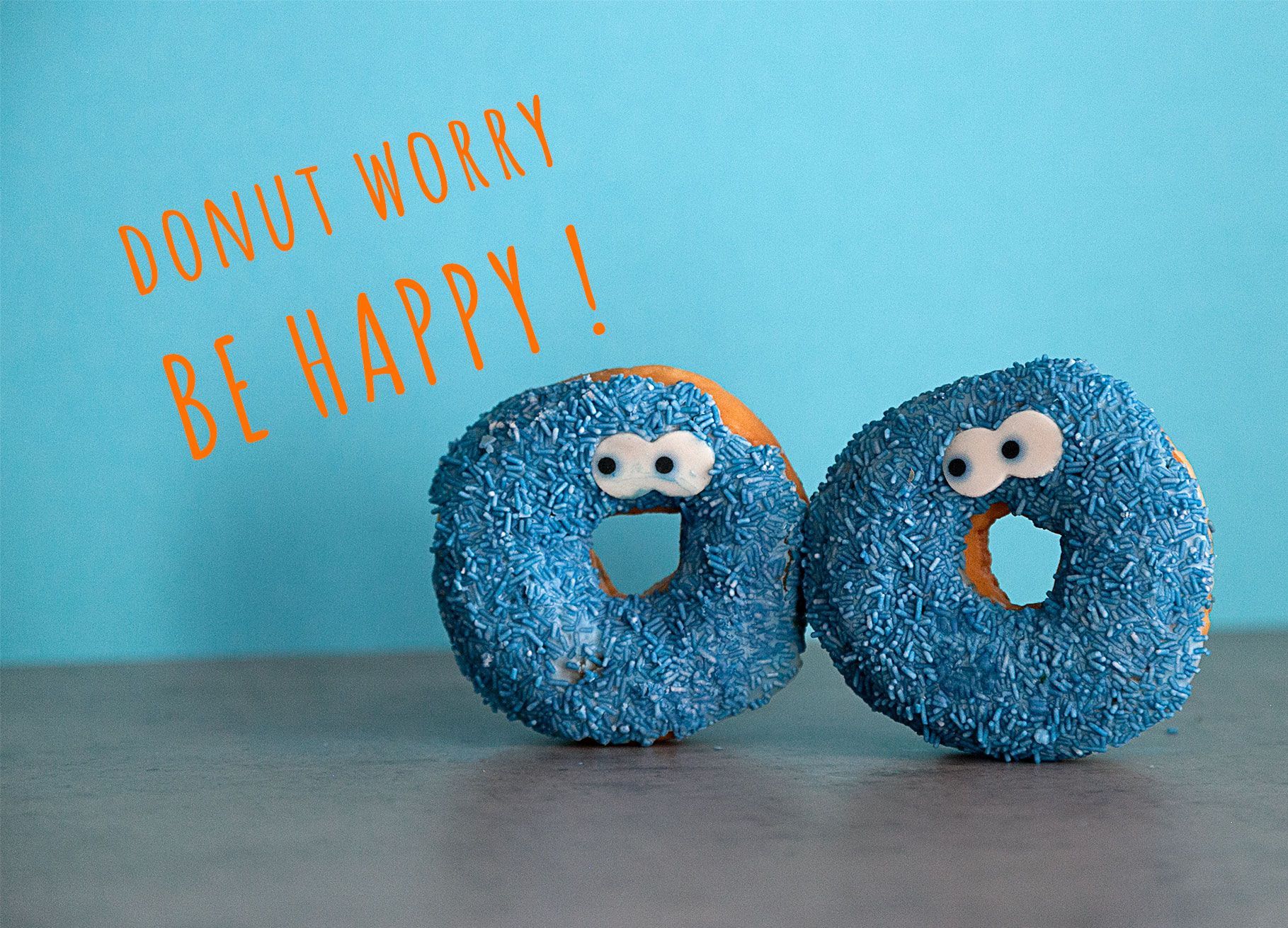 Zwei Donuts mit blauen Zuckerstreuseln und Augen stehen vor einem hellblauen Hintergrund. Daneben steht auf der Postkarte der Spruch: Donut worry. Be happy!