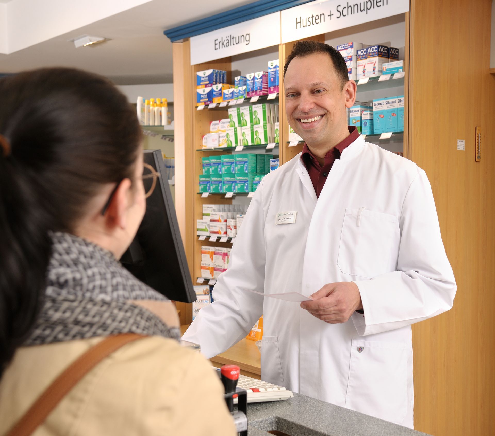 Apotheker Markus Titzek berät Kundin in der Süd Apotheke Gütersloh am HV-Tisch vor dem Regal mit Erkältungs- und Hustenmitteln.