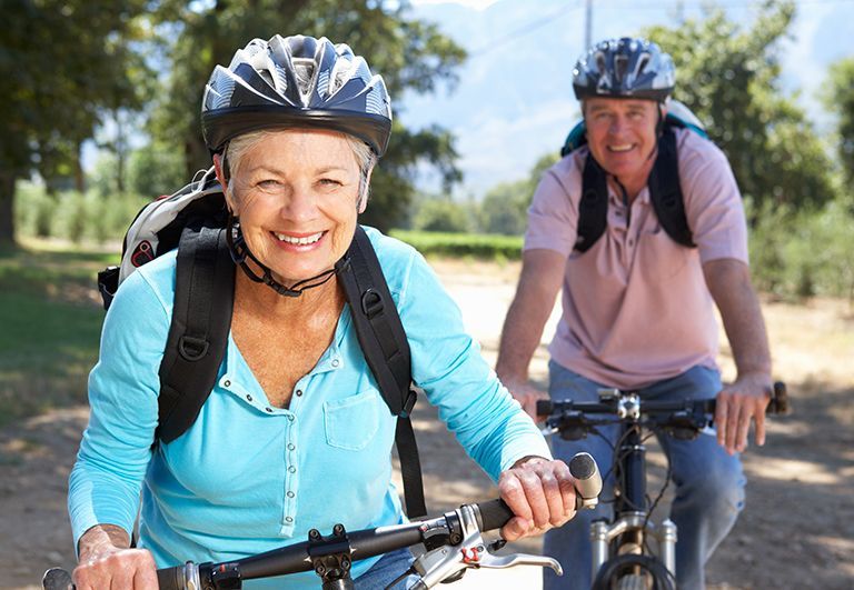 Im Vordergrund eine ältere Dame mit Fahhradhelm und türkisfarbenem Shirt auf Fahrrad, im Hinetrgrund Mann mit Helm und rosa Shirt