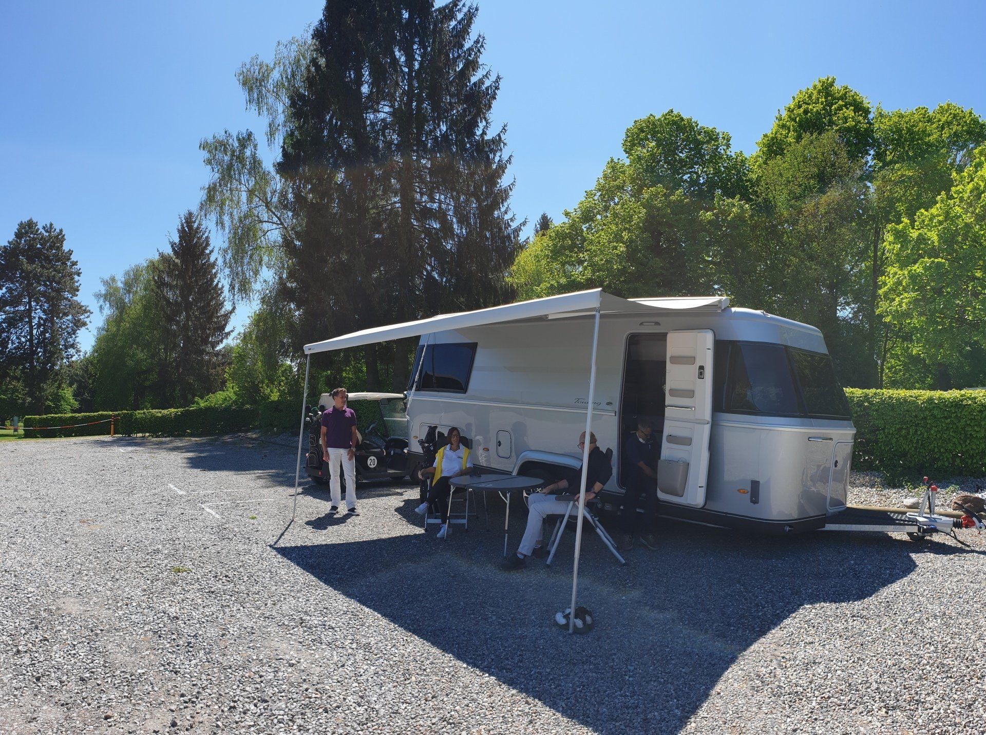 Wohnwagen auf Stellplatz mit Personen sitzend im Vordergrund