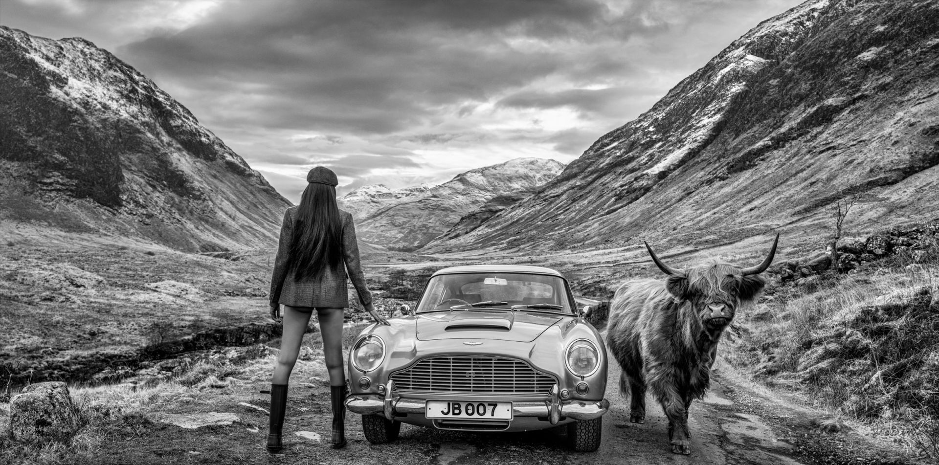 Black and white photograph of James Bond Aston Martin with female model and highland cow in Glencoe Scotland by David Yarrow