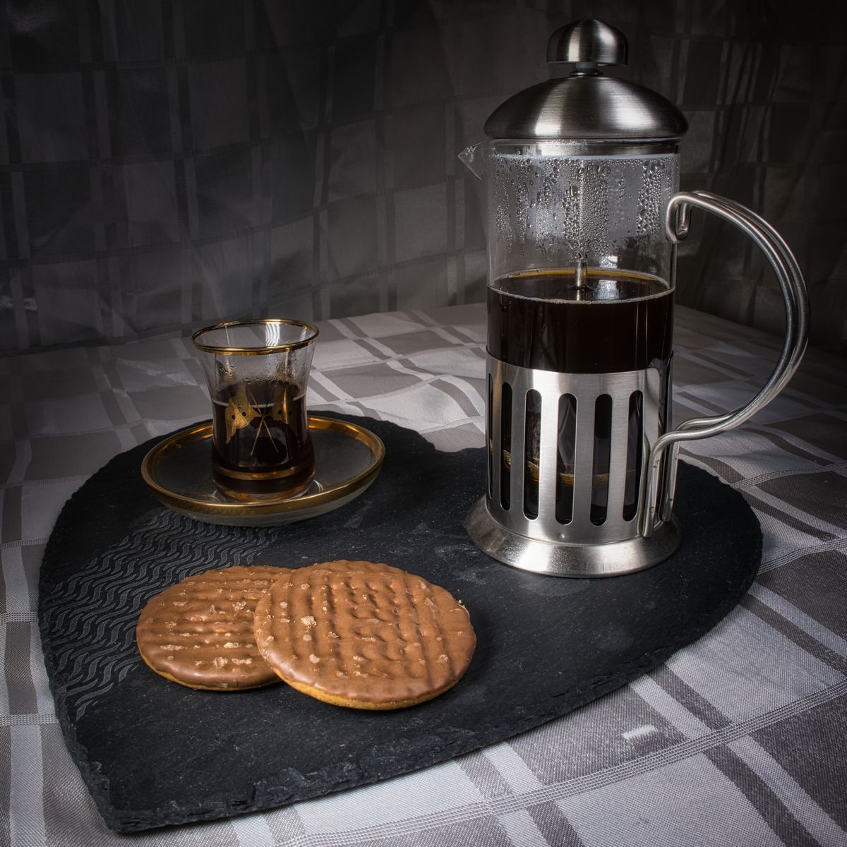 A still life image of a coffee cafetiere, Turkish tea glass, and two chocolate biscuits