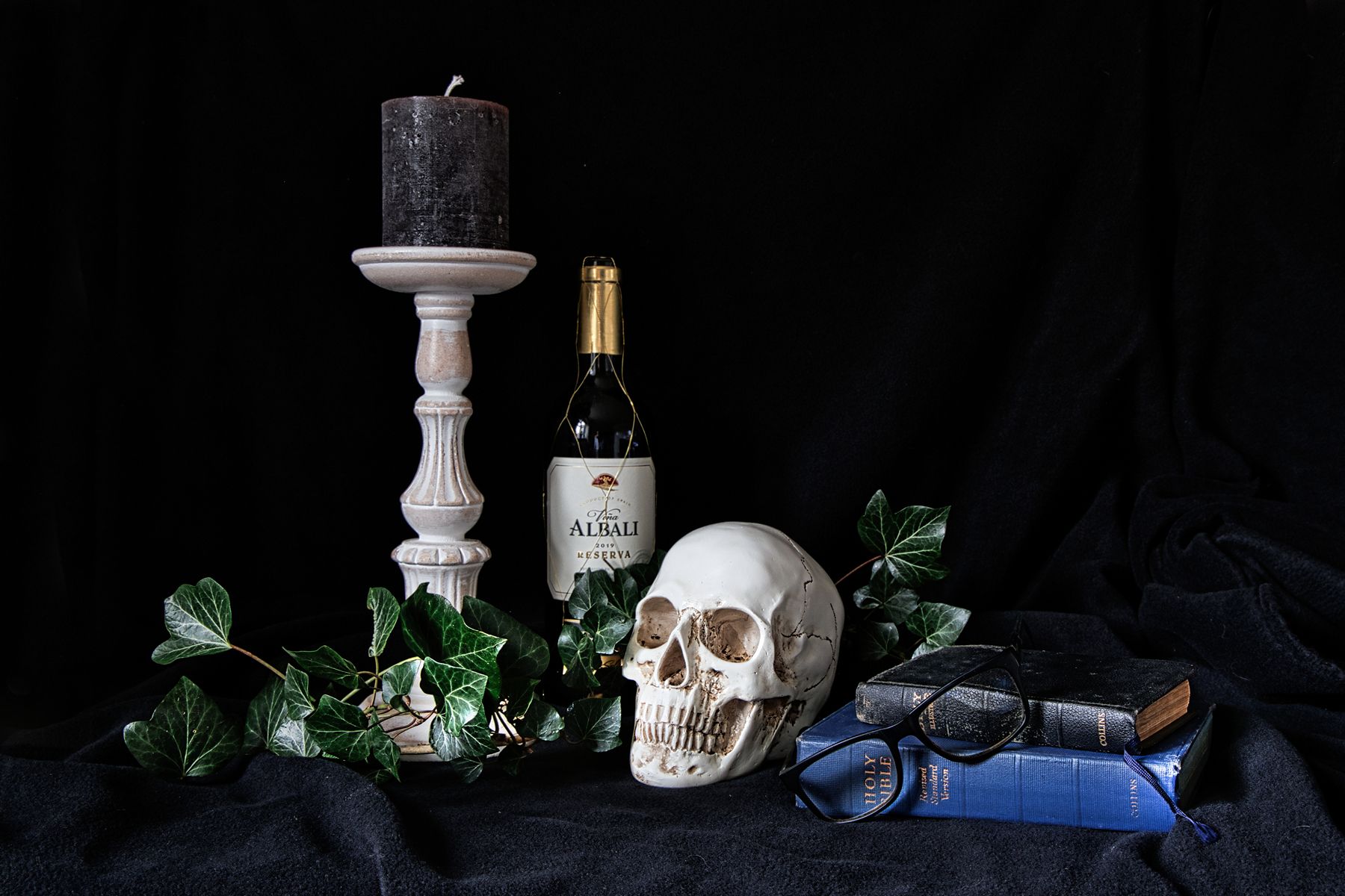 A still life image of a black candle, on a white candle stick, a bottle of port, a human skull, and two Bibles