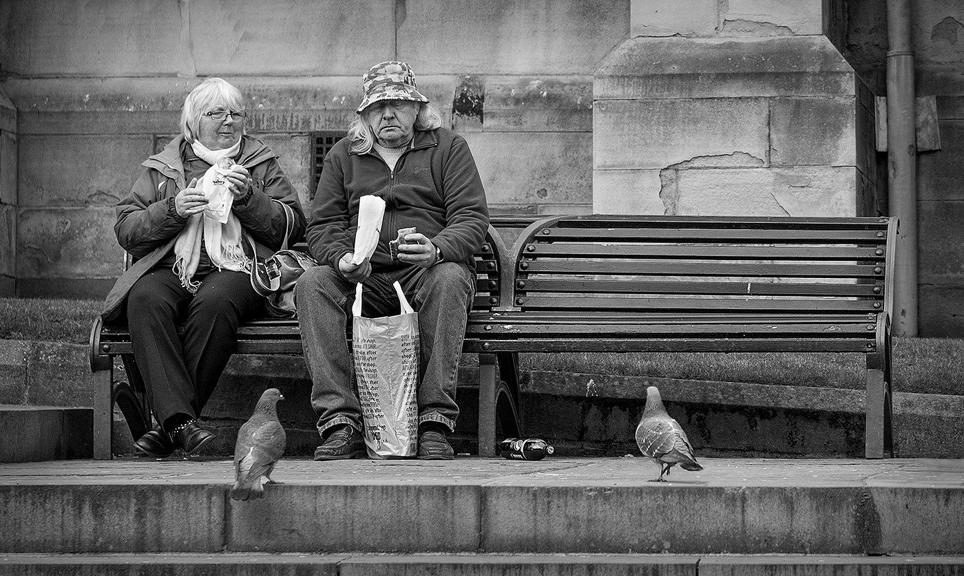 a couple seated on a bench eating lunch, with pigeons at their feet