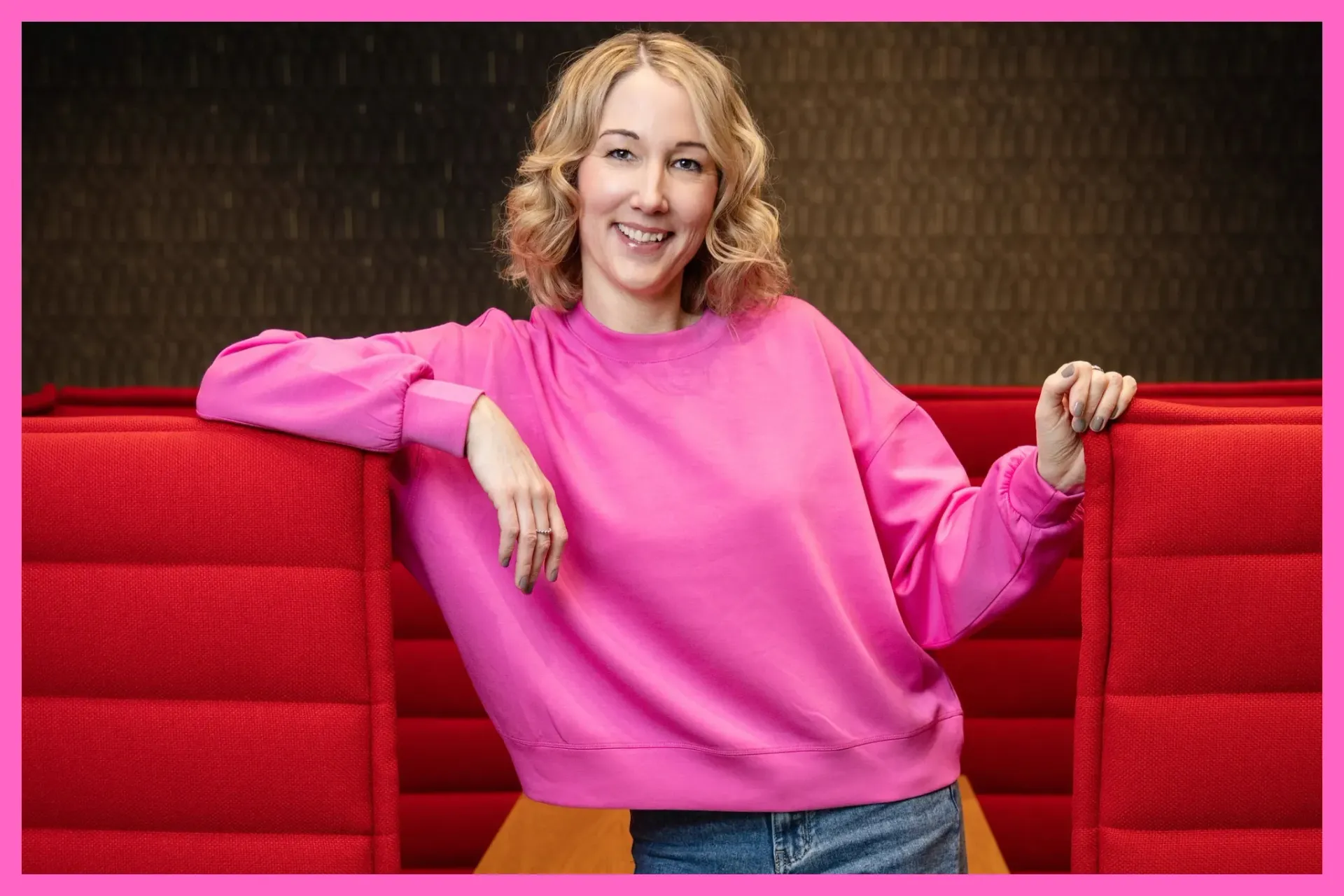 Corine Brötz steht lächelnd vor einer roten Akustikwand und trägt einen pinkfarbenen Pulli und eine Jeans.