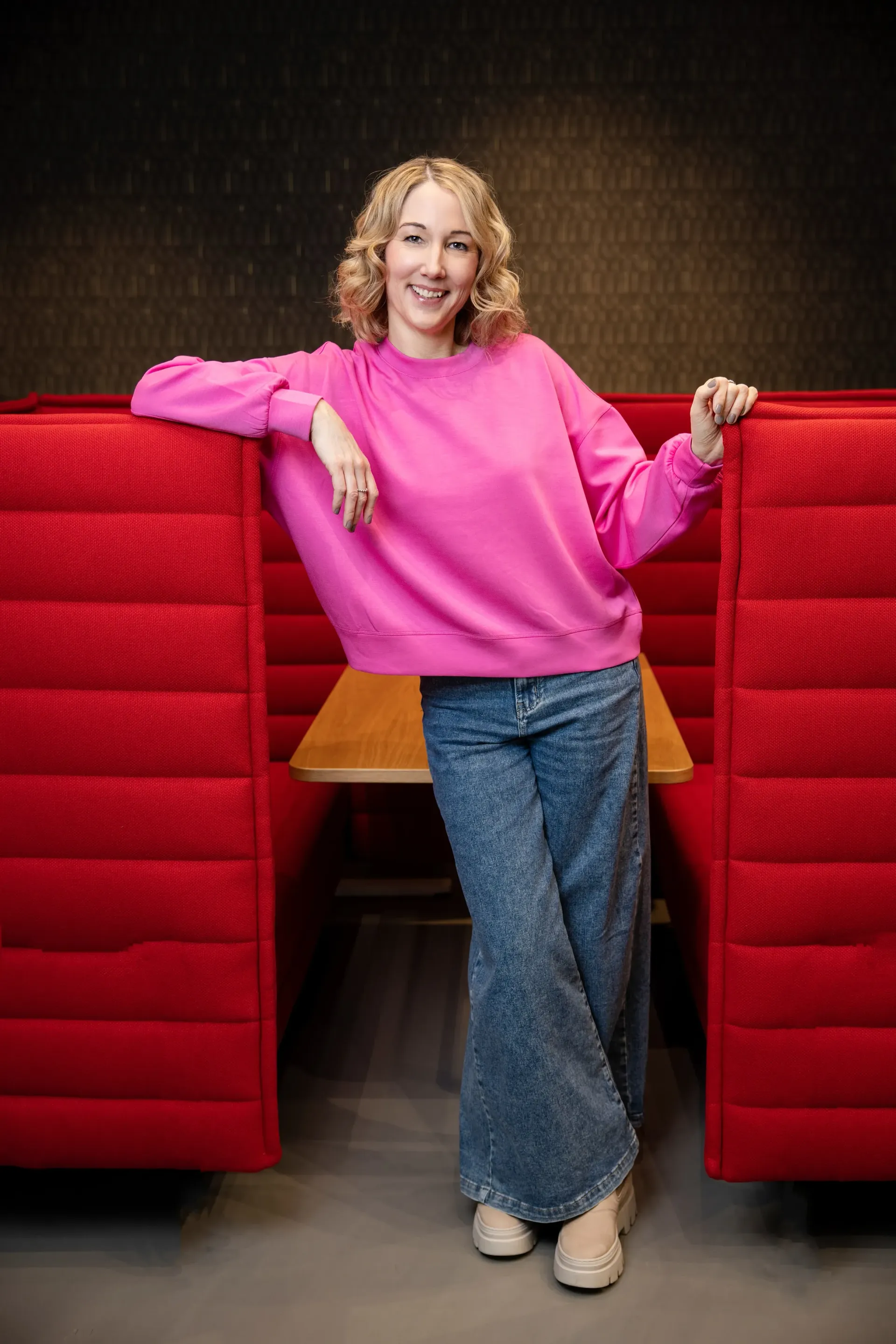 Corine Brötz steht lächelnd vor einer roten Akustikwand und trägt einen pinkfarbenen Pulli und eine Jeans.