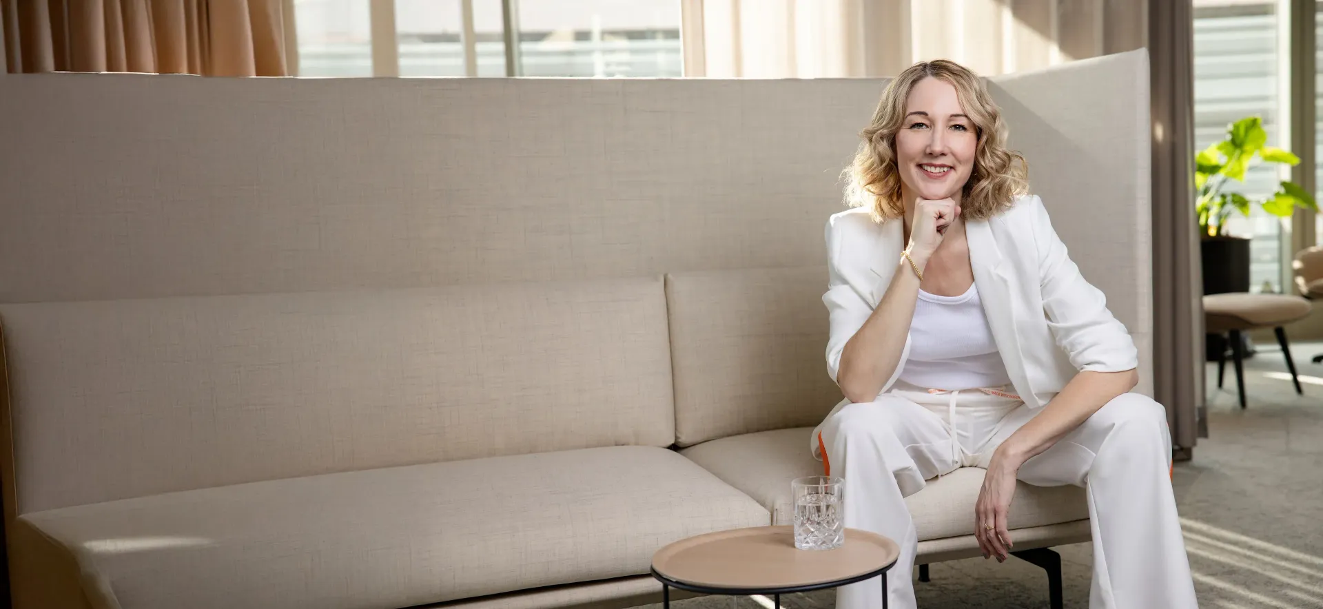 Corine Brötz – Sprecherin
Sie sitzt in einem weißen Hosenanzug auf einem beigen Sofa.