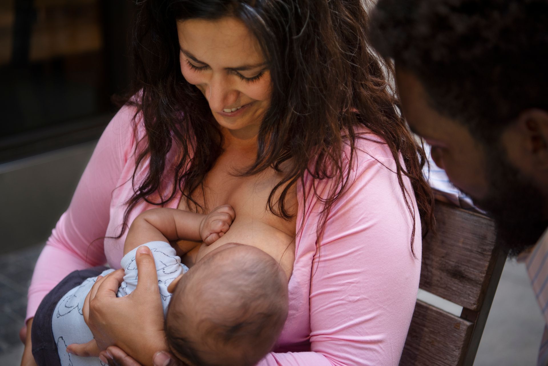 A woman with dark hair, wearing a pink, unbuttoned blouse, is sat on a wooden seat looking down smiling at her breastfeeding baby.  A man with dark hair and a beard looks on from one side smiling.  The baby is dressed in a shirt and trousers and is viewed from behind as he feeds.
Designed by freepik