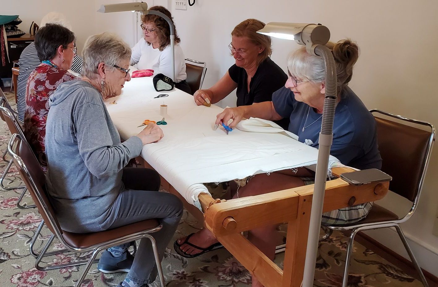A group of women sit at a long quilt frame appearing to talk while hand quilting.