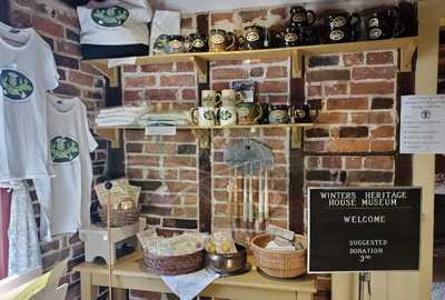 Image of an old but neat brick wall with t-shirts, tote bags, and stoneware with the Winters Heritage House pipes and thistles logo plus baskets with colonial-style toys displayed on an entryway table