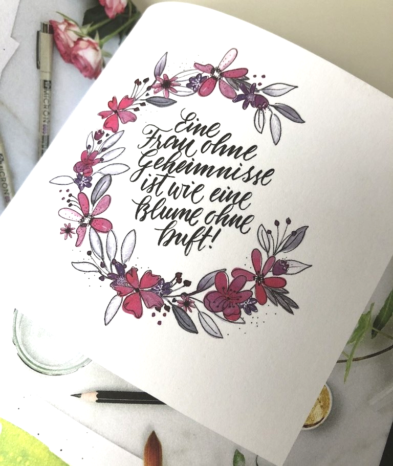 Handlettering in moderner Kalligrafie in schwarz. Das Handlettering ist von einem Blumenkrank aus Watercolorblumen umschlossen. Die Blumen sind lila und pink.