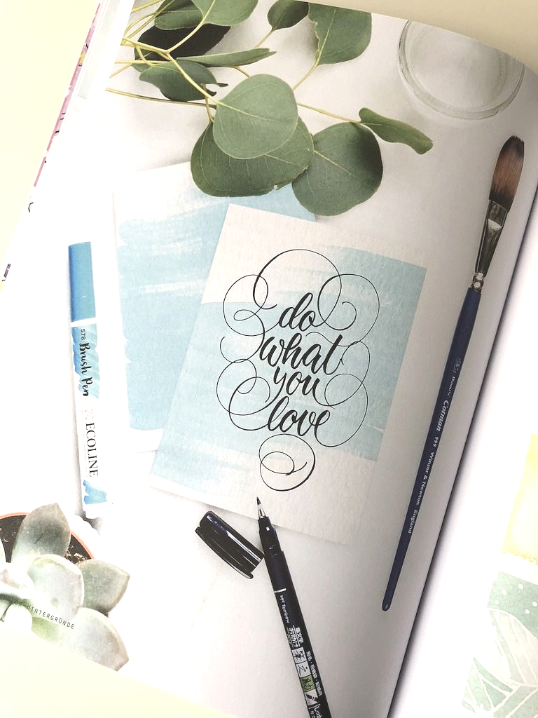 Schwarzes, handgeschriebenes Lettering in Schnörkel-Schreibschrift auf hellblauem Watercolor- (Aquarell) Hintergund.