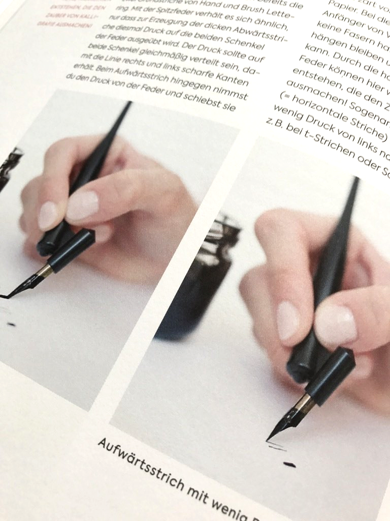 Handlettering. Frau Hölle zeigt in ihrem Buch Handlettering-Alphabete, wie sie mit einer Kalligrafie-Feder schreibt. Das Bild zeigt die Feder in den Fingern, die mit Tinte aus einem Tintenfass befüllt ist.