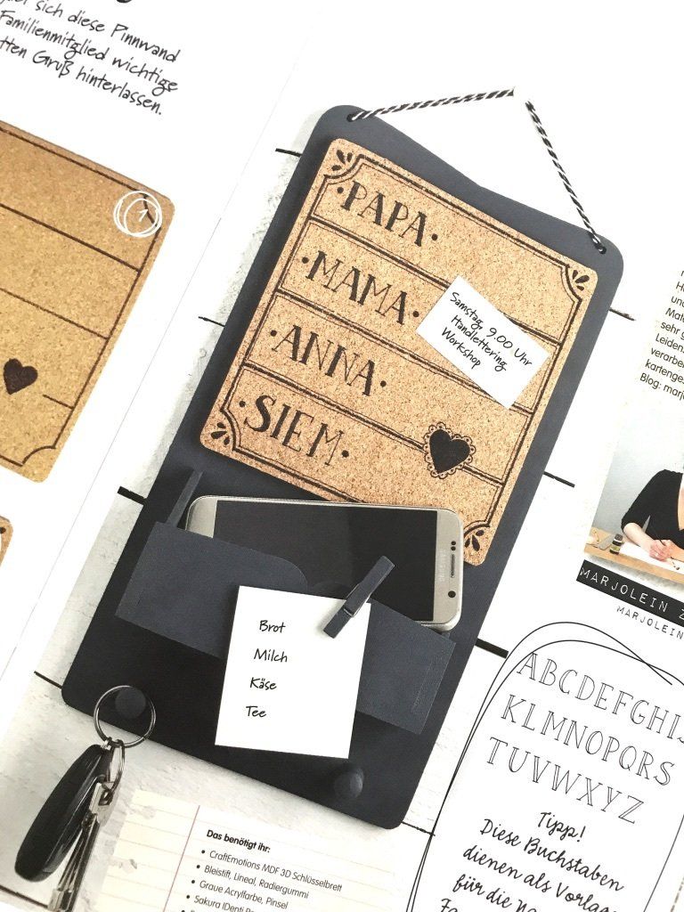 DIY-Schlüsselbrett in schwarz/grau. Einzelne Haken mit Schlüssel, Tasche fürs Handy, Zettel mit Wäscheklammer angeheftet und viereckiges Korkschild von Hand mit Mama, Papa, Anna und Siem belettert.