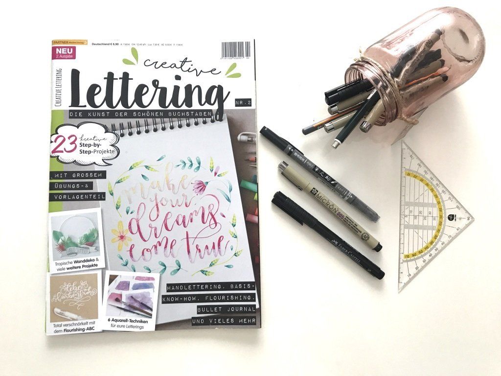 Das Magazin creative Lettering liegt auf einem weißen Tisch. Daneben liegt ein rosafarbenes Deko-Glas, das mit Fineliner, Pigmentmarker und Brushpens gefüllt ist. Daneben liegt ein Geo-Dreieck.
