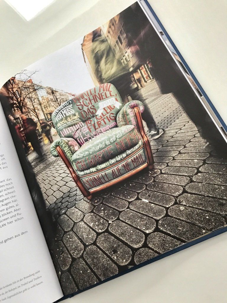 Ein altmodischer Ohrensessel steht auf Kopfsteinpflaster in einer Fußgängerzone. Dieser Sessel ist von Hand großflächig beschriftet.