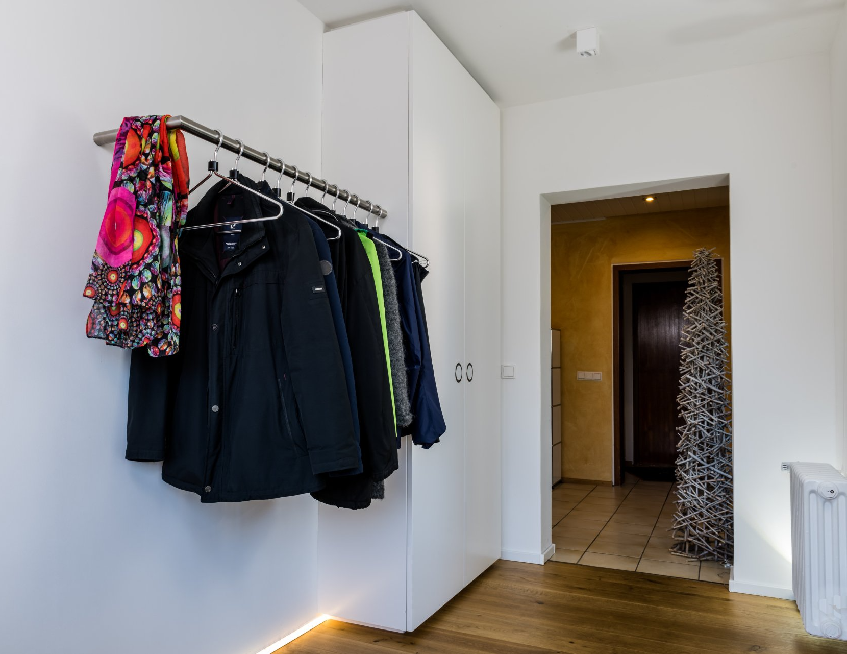 Garderobe von Schreinerei BLEIER Lebensräume, Garderobenstange mit Haken und Kleidung, dahinter ein hoher weißer Schuhschrank