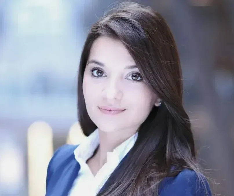 Lächelnde dunkelhaarige Frau mit langen Haaren, dunkelblauer Blazer, verschwommener Hintergrund