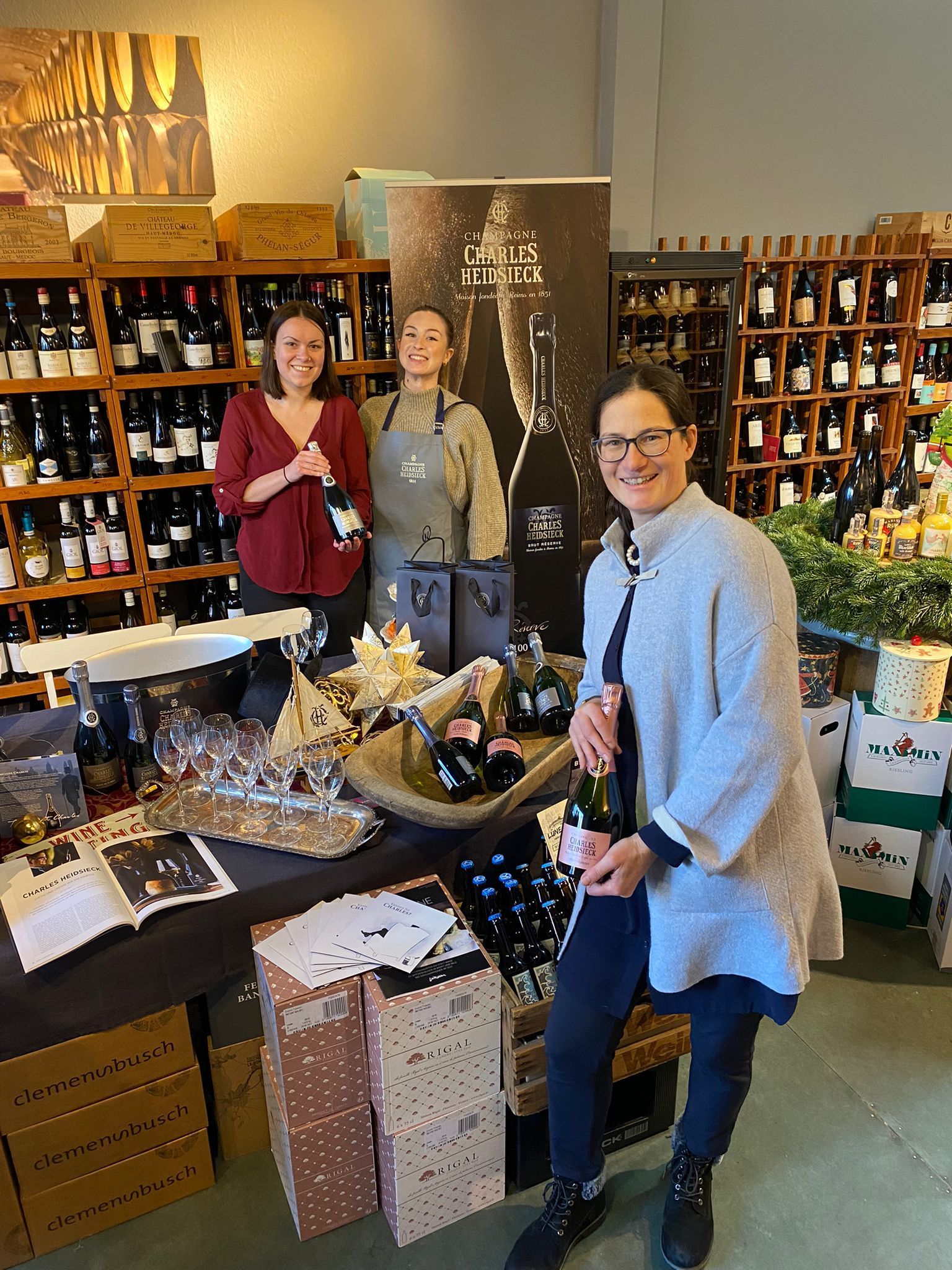 Drei Frauen stehen an einem mit Champagnerflaschen und Gläsern bedeckten Tisch und blicken lächelnd in die Kamera.