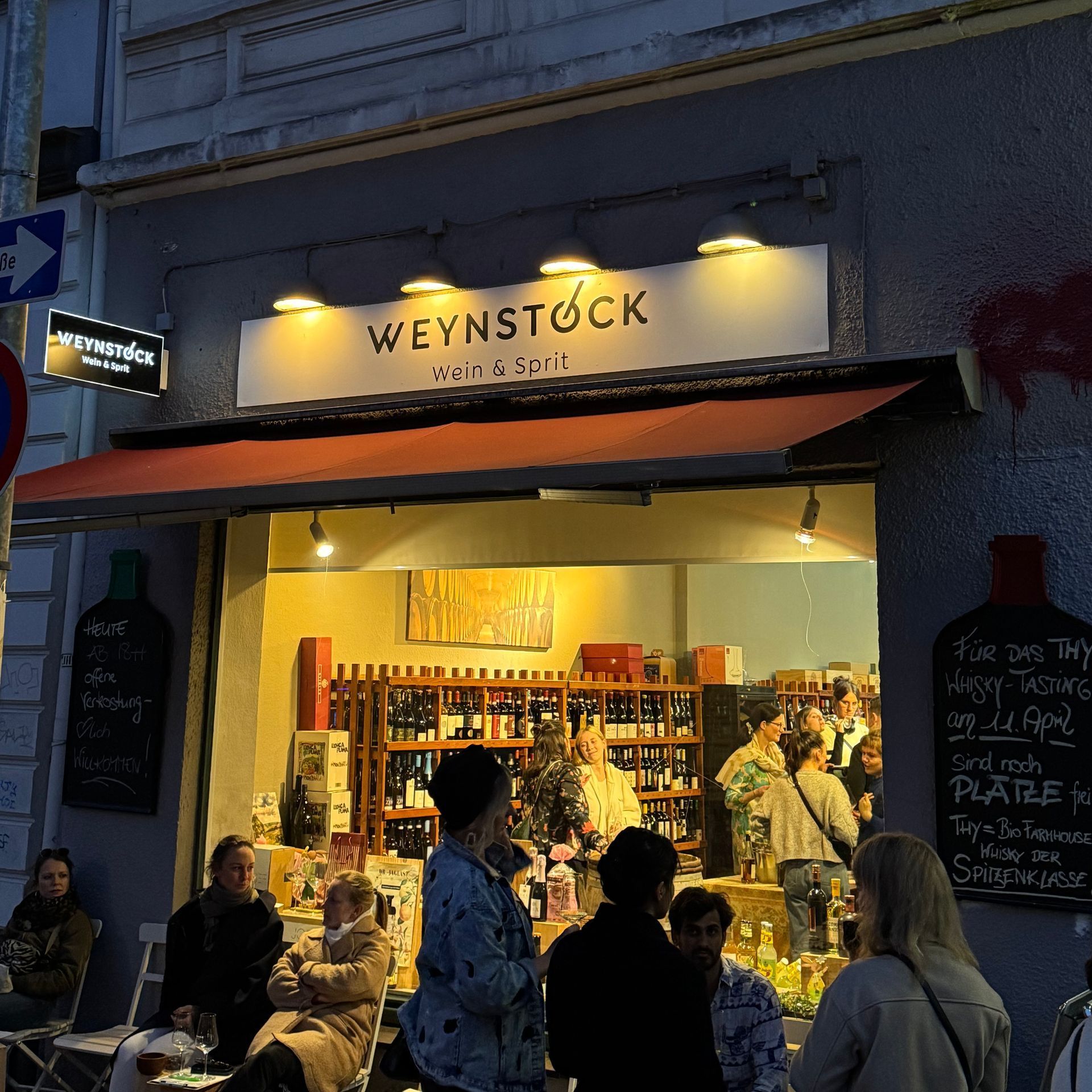 Ein volles Weingeschäft wird von draußen gezeigt, vor dem Geschäft sitzen Personen auf Stühlen.