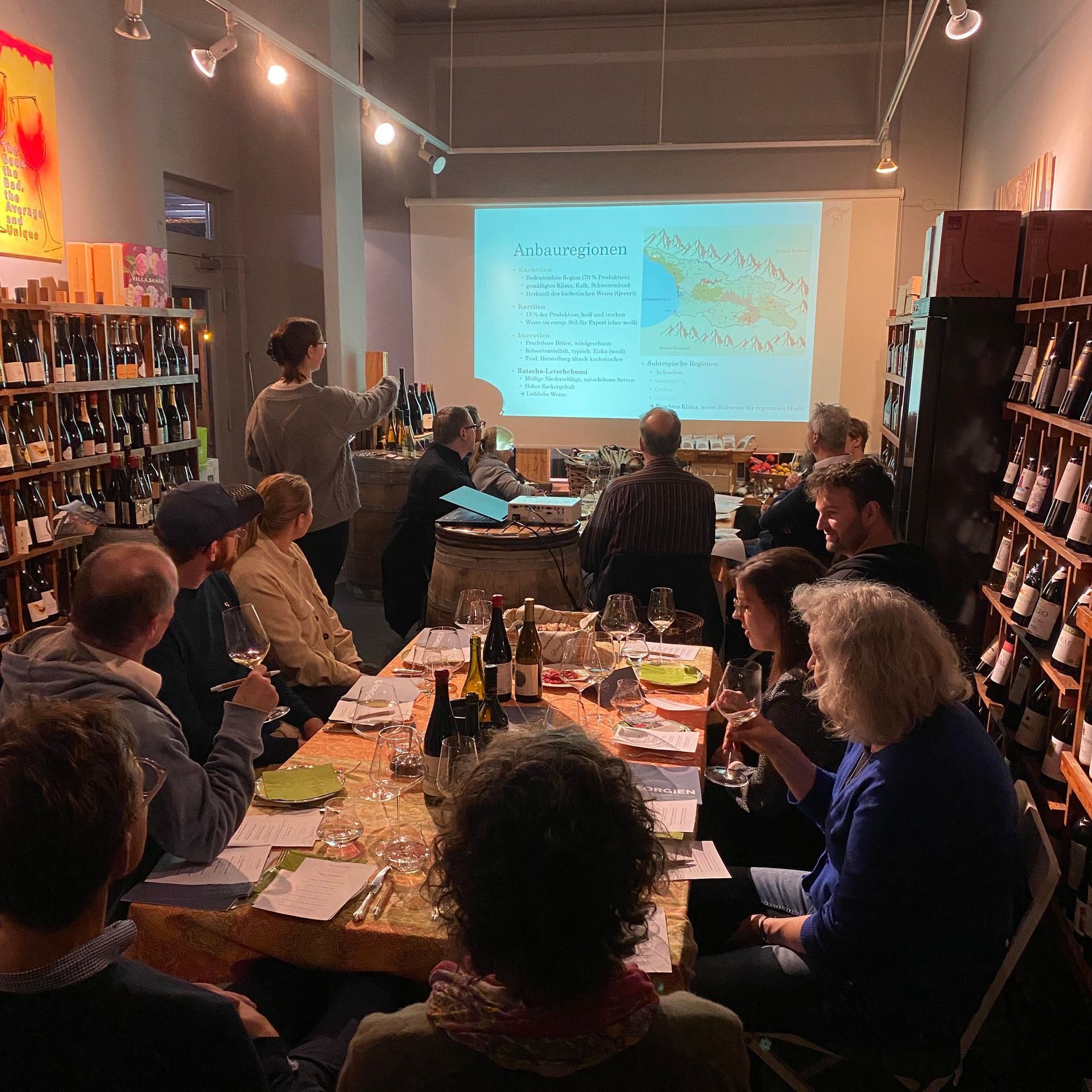 Ein Dutzend Menschen sitzen an zwei gedeckten Tischen und blicken an die Wand, auf die ein Bild projiziert ist.