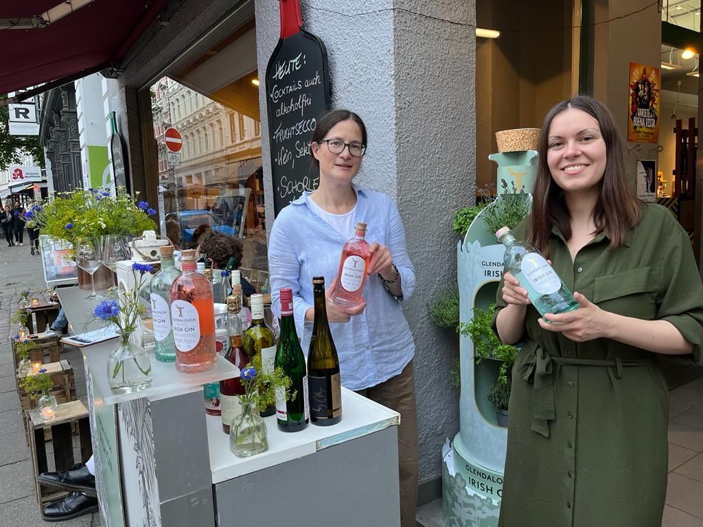 Zwei Frauen stehen hinter einem auf der Straße aufgebauten Weinstand und lächeln in die Kamera.
