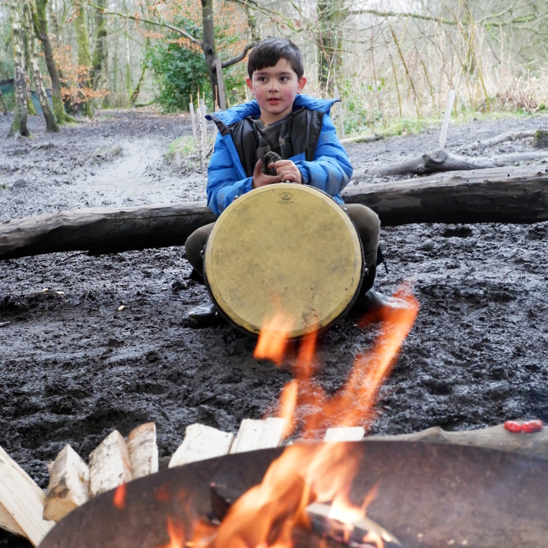 Boy at Summerseat holiday club playing in the drums sat on a log in front of the firepit