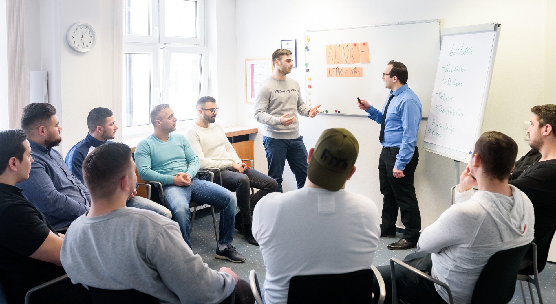 Unterrichtsszene vom AEVO-Kurs bei ASM Eine Gruppe von Männern sitzt im Stuhlkreis. Ein Teilnehmer steht und interagiert mit dem Dozenten.