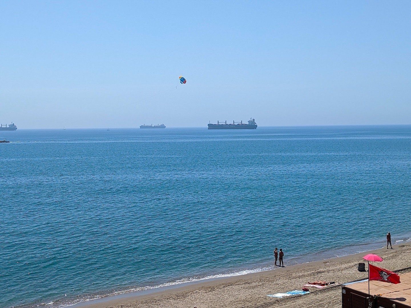 Mulitcoloured parachute in the air at sea with ships and people standing on the shoreline